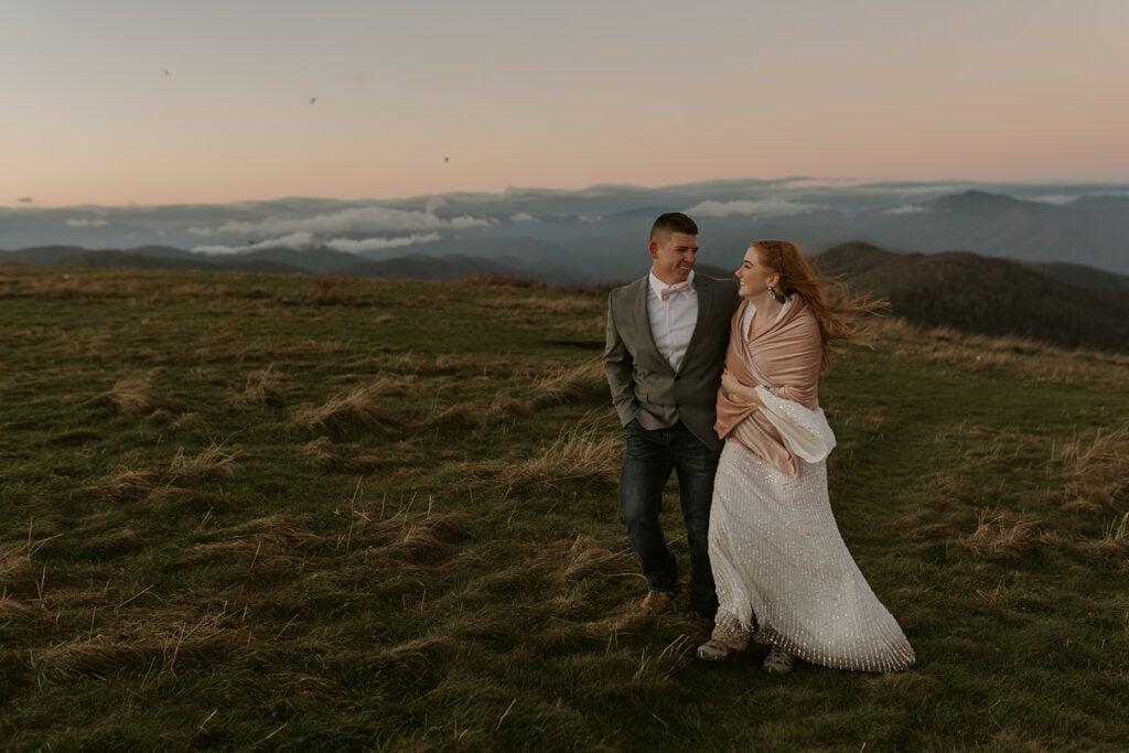 Two people are walking on a mountaintop at Max Patch during sunrise looking at each other and smiling. They are in their elopement outfits of a dress and suit.
