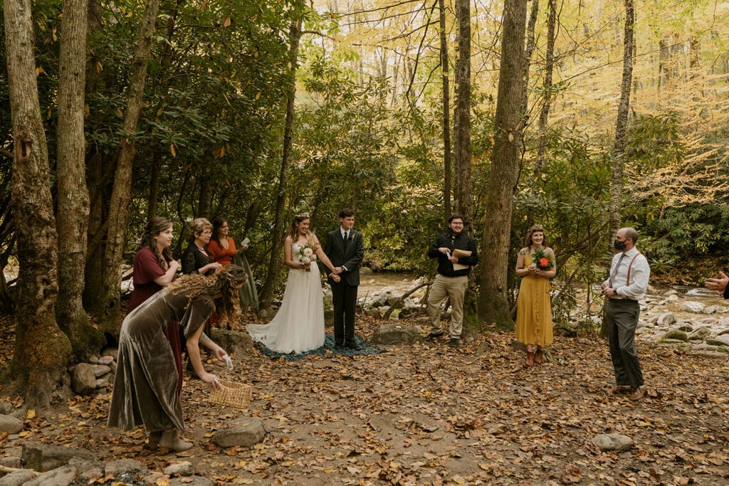 A ceremony of a small wedding in the woods with fall colors and a river in the background.