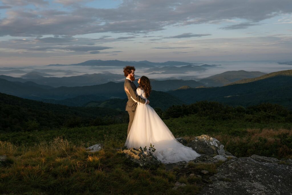 A couple stands in North Carolina, one of the best places to elope, on their wedding day hugging and looking at each other in front of a cloud inversion mountain landscape.