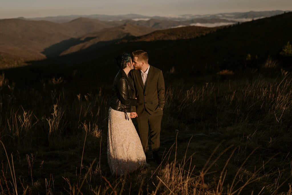 A couple stands on a mountain in their wedding clothes. The groom is kissing the bride on the cheeck.