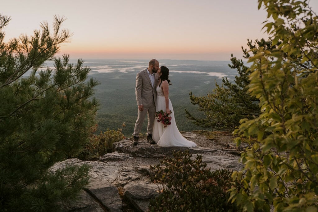 A couple stands kissing on a rock formation surrounded by trees and blue mountains for their elopement.