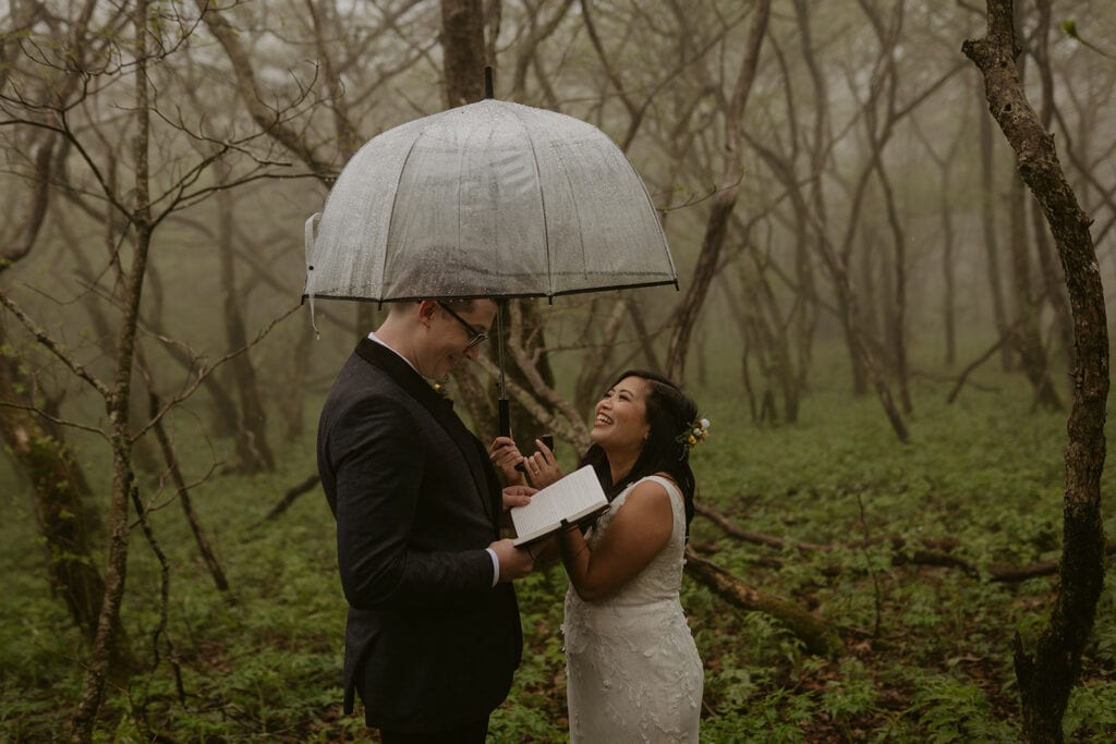 A couple stands in a foggy forest with an umbrella over them. The groom is reading from a book and the bride is smiling up at him.