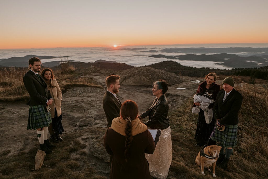 A couple is having a sunrise ceremony on Black Balsam knob for their wedding. The couple stands in front of an officiant with two people on either side of them. The mountains are blue in the background with a cloud inversion.