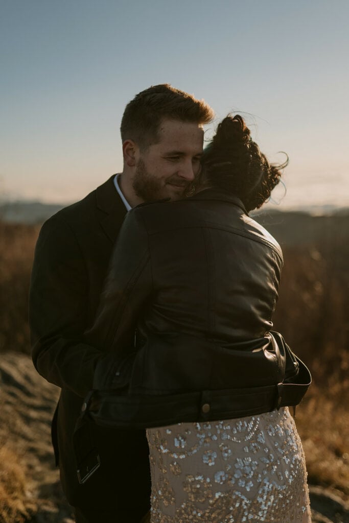 A couple stands hugging in warm golden light on a mountain top. We can see the face of the man who is smiling and his hands are wrapped under the woman's leather jacket. She has a sparkly dress on underneath.