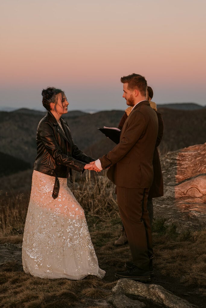 A couple is standing holding hands at sunrise smiling at each other during their black balsam knob wedding ceremony. The sky is pink and there are mountains behind them.