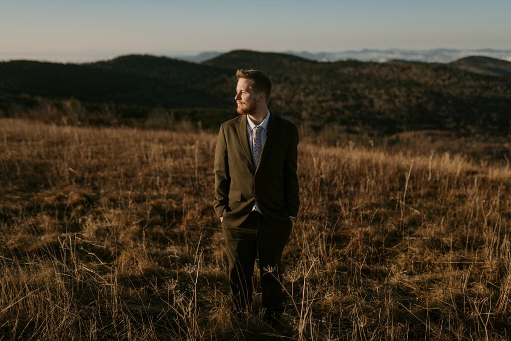 A groom is standing in front of a landscape in his wedding clothes with mountains behind him.