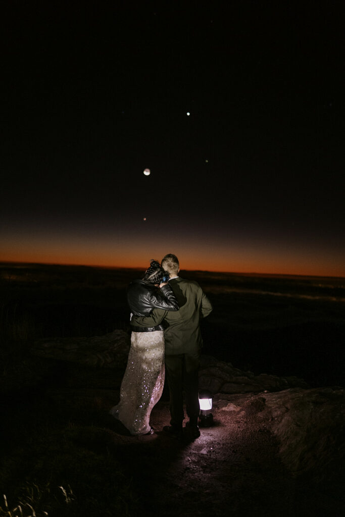 A couple stand with their back to the camera looking out over a landscape at night with an orange light coming over the horizon. There are stars and the moon in the sky. They are dressed in wedding clothes and beside a lantern on the ground.