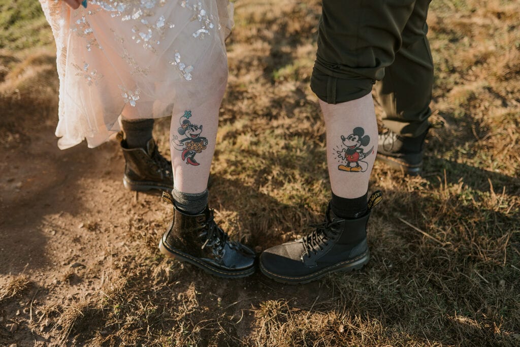 A couple is showing off their matching Mickey and Minnie Mouse tattoos on their calves. The girl lifts up her dress and the guy lifts up his pant leg. They have matching doc martins on and are standing in golden grass.