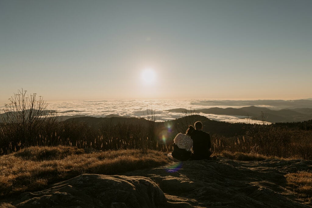 A couple sits snuggled up facing out at a view not long after sunrise at Black Balsam Knob after their wedding. The sun is bright and illuminates a cloud inversion