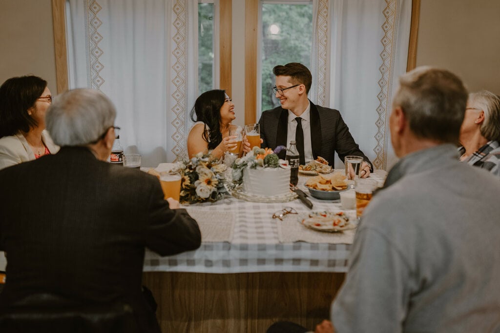 A couple and their family are sitting around a table holding beers up for their elopement. There is a cake in front of them and they are smiling at each other.