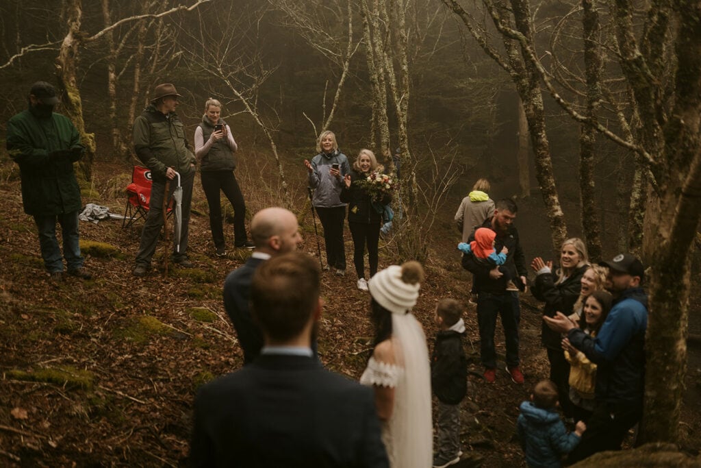 A family stand around cheering for a couple that just ended their elopement ceremony in the foggy woods.