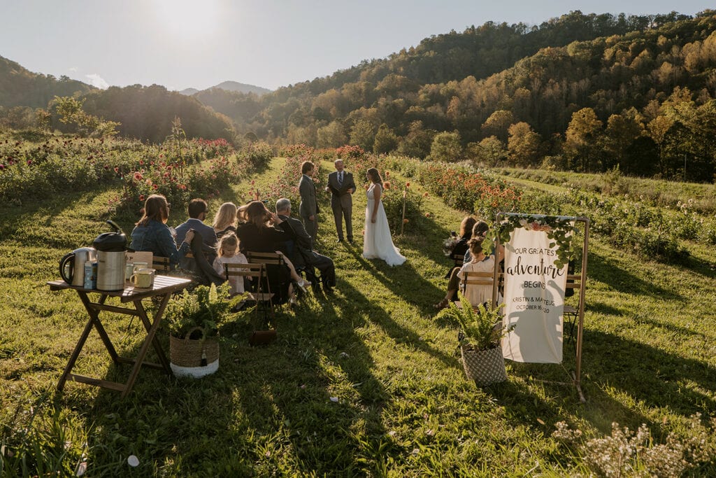 A small ceremony is happening in a flower farm in NC right before sunset with bright sunshine. The couple is standing with the officiant as the family sits and watches.