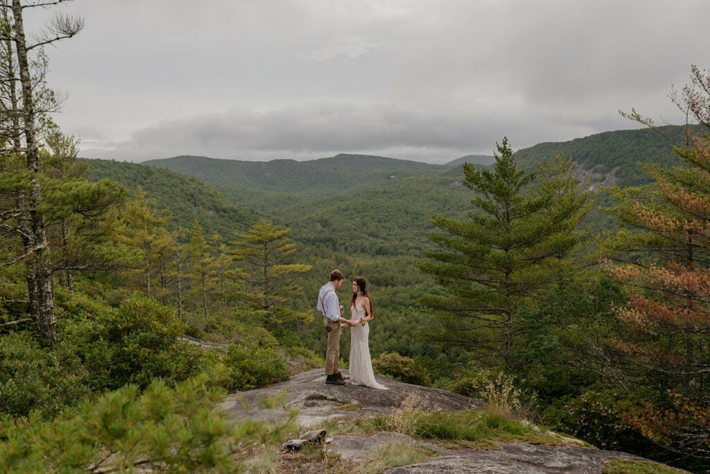 A couples stands in their wedding clothing facing each other with a summer mountain view and forest behind them.