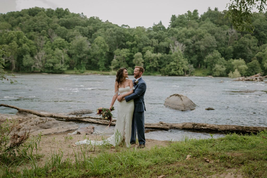 A couple stands hugging in front of a beautiful wide river in their wedding clothes. They are smiling at each other and the bride is holding a bouquet.