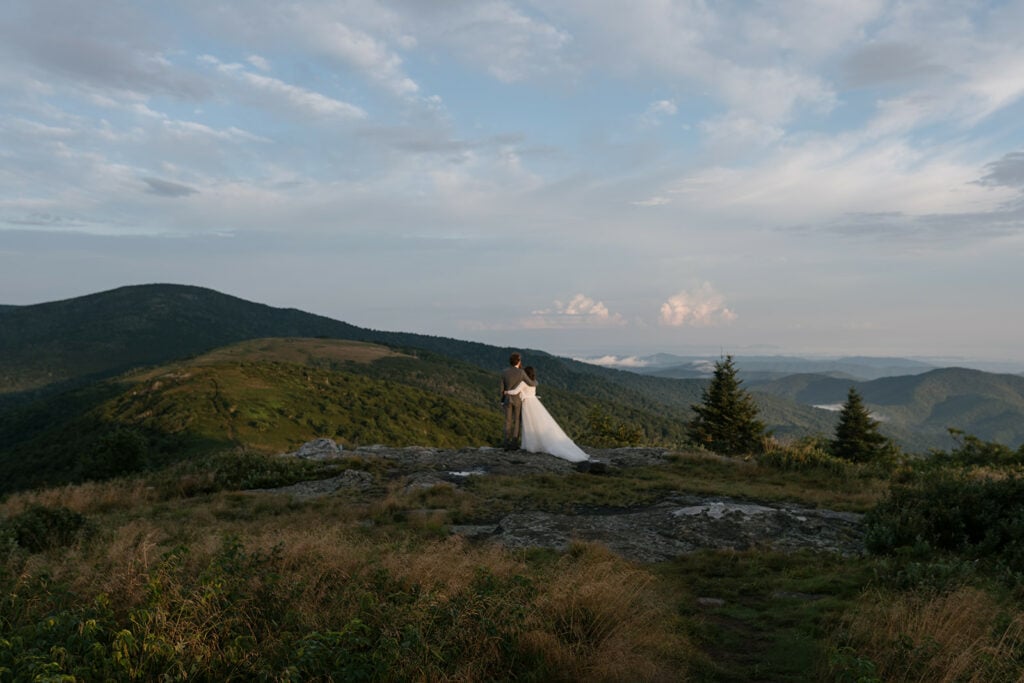 A couple is standing in their wedding clothes, hugging, looking out at the view on Roan Mountain, one of the best places to elope in NC.