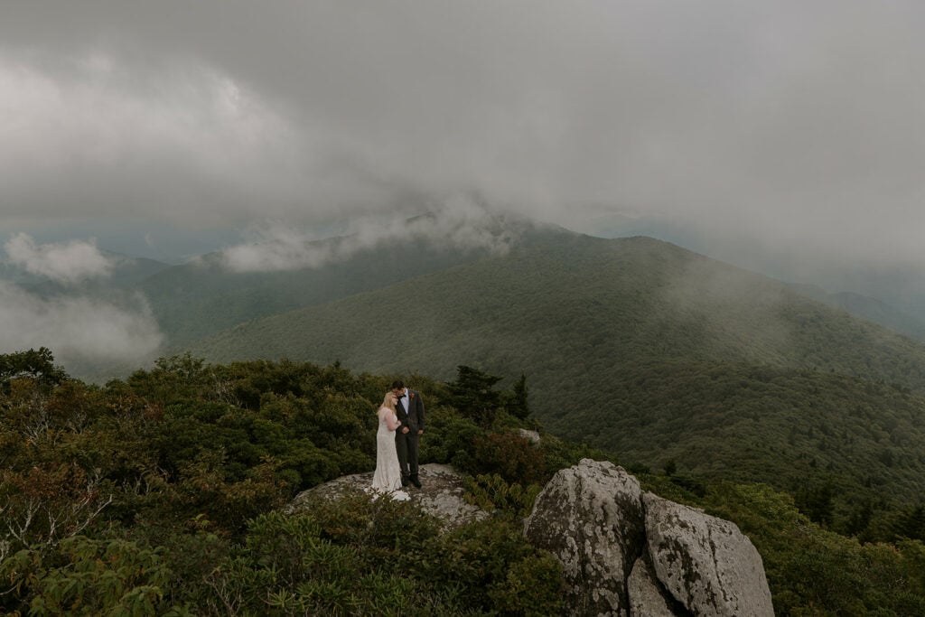 A couple is standing in their wedding clothes, hugging, looking out at the Blue Ridge mountain views, one of the best places to elope in NC.
