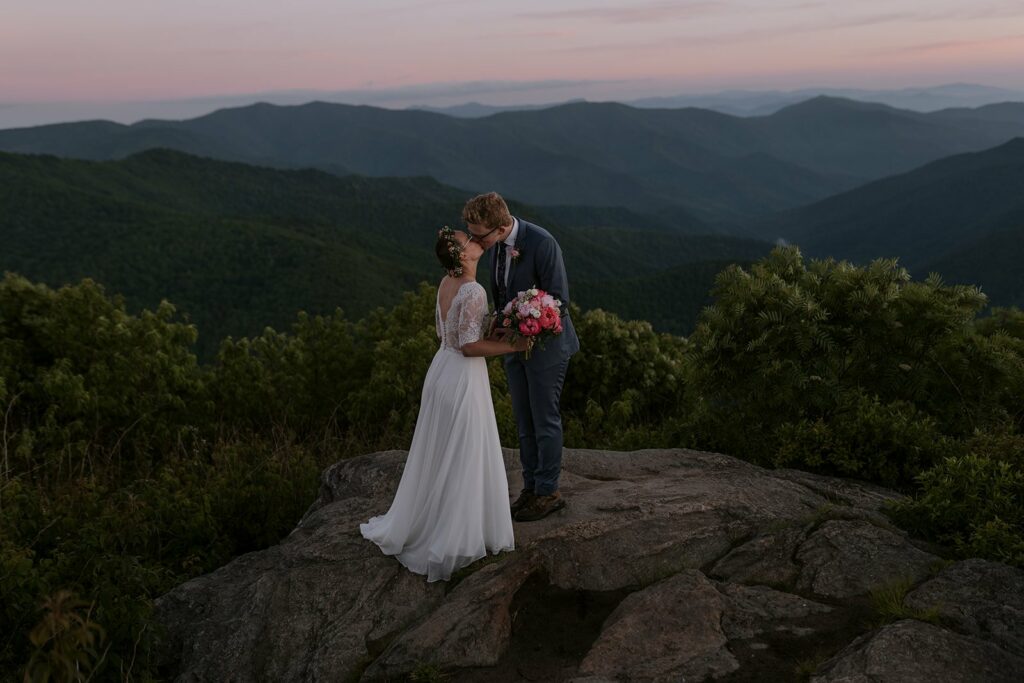 A couple stands on a mountain at sunrise kissing during their Asheville elopement.
