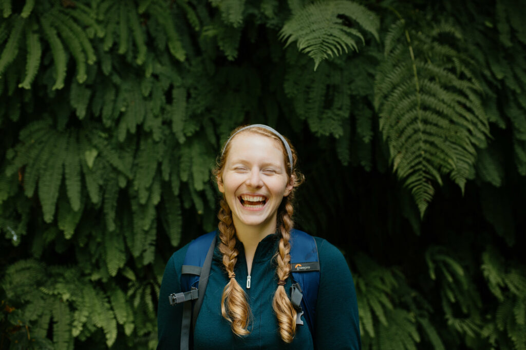 A redheaded girl stands in front of a wall of ferns with her hair in braids smiling with her eyes closed. She has a blue shirt and backpack on with her hair in braids.