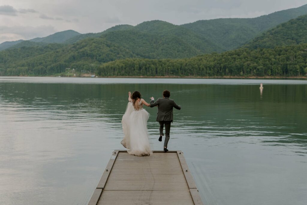 A couple is jumping in the lake for their Asheville elopement. They are in their wedding clothes surrounded by mountains and jumping off the dock into the water while holding hands.