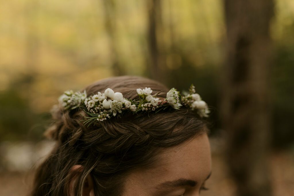 A close up shot of a flower crown atop a brunettes head. She is in a yellow fall forest and her crown is made of greenery and little white florals and berries.