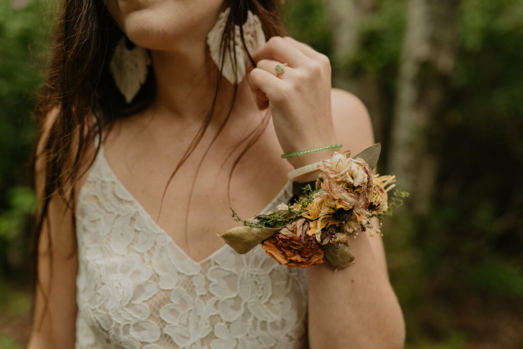 A close up shot of a bride on her elopement day with a floral wristlet made of dried flowers for her wedding. She has a lace dress on, long brown hair, macrame earrings, and is in the forest.