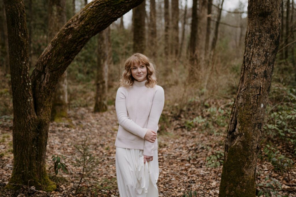 A bride is looking away from the camera in her elopement outfit (a soft light colored sweater and white skirt) and is standing in a winter forest.