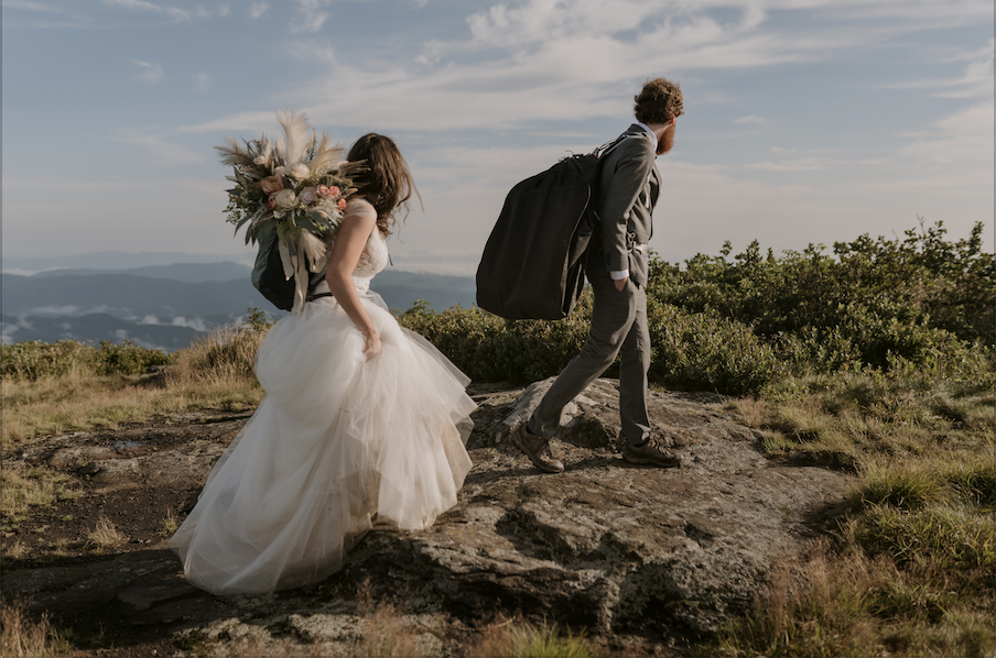 A couple begins a hike on their elopement day in their wedding clothes looking away from the camera out at the mountains. The bride has a beautiful rental bouquet in her backpack.