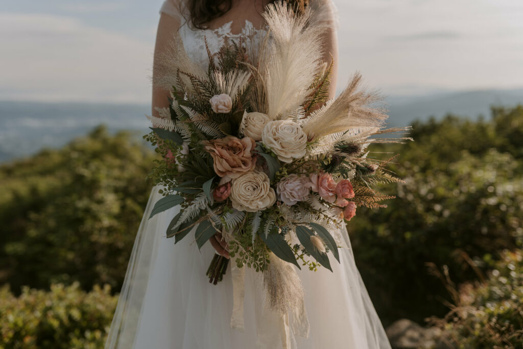 A close up of a bouquet being held by a bride on a mountaintop with a view in the background. The flowers are made of silk and are green, pink, and beige.