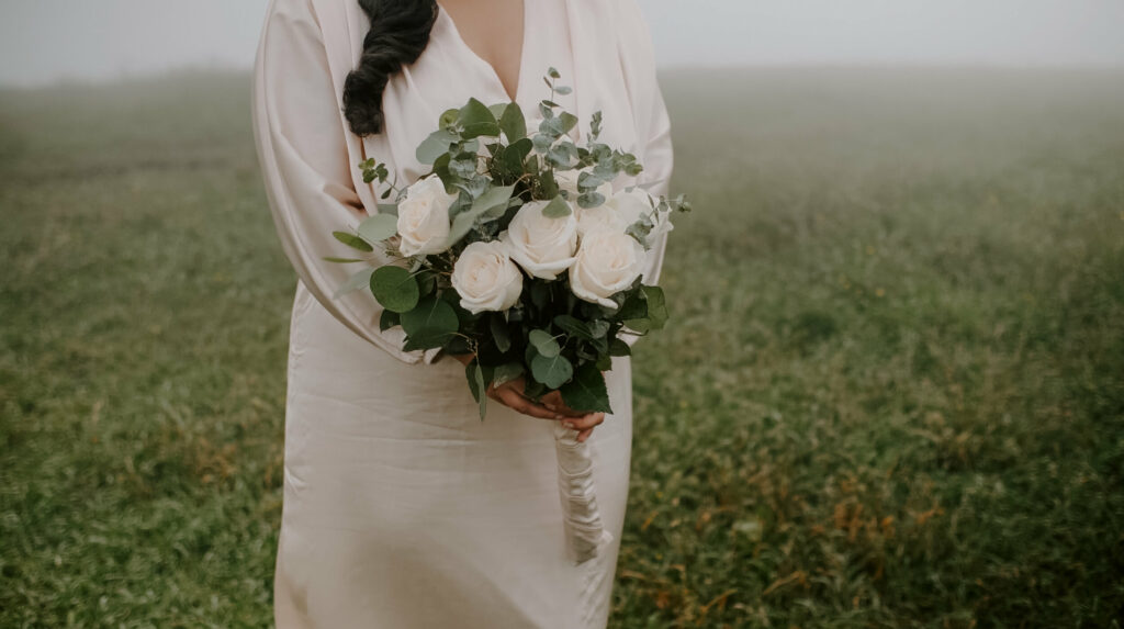 Someone is holding a traditional roses and eucalyptus bouquet on a foggy mountaintop in a beige silk dress.