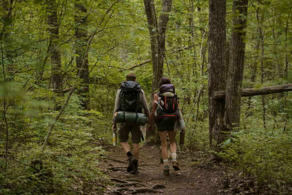 A couple is holding hands and hiking through the woods, backpacks full of gear for their camping elopement in North Carolina.