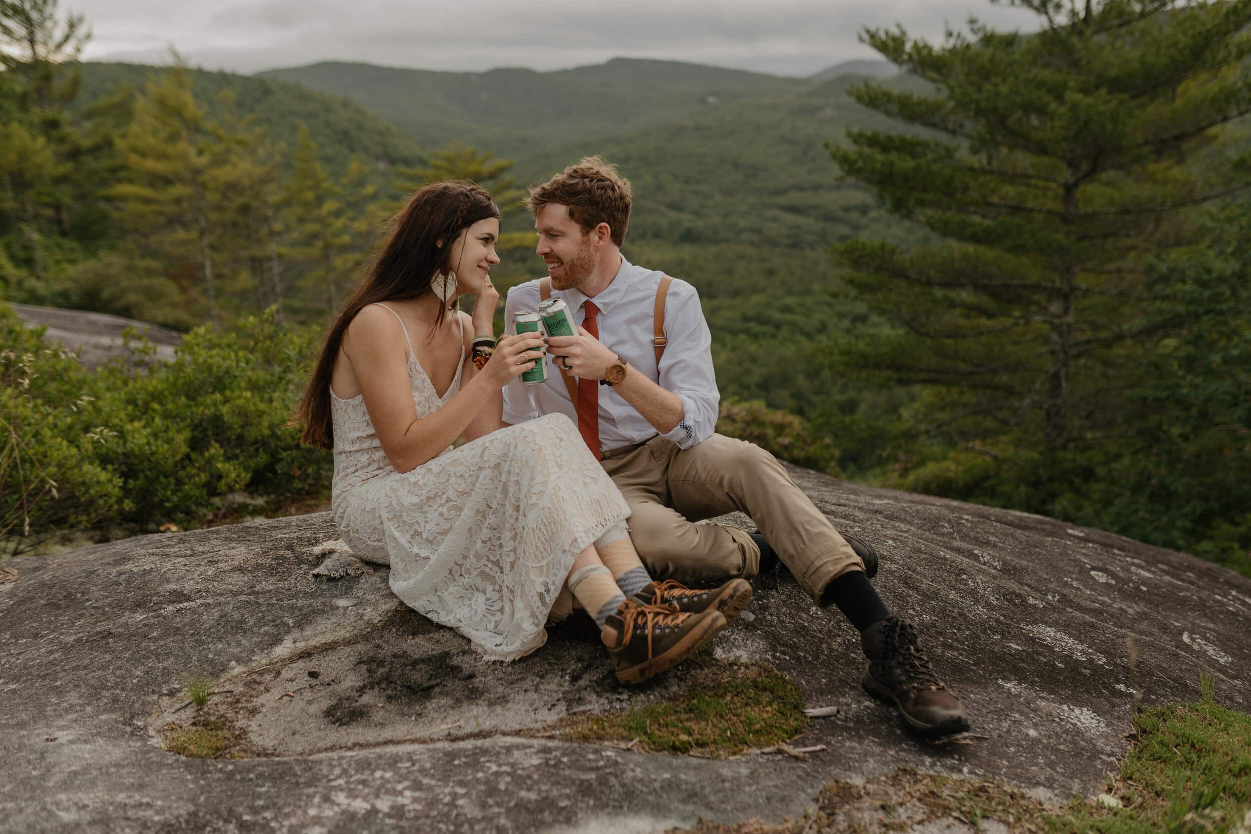 A couple is holding canned drinks, sitting on a rock while they elope in Asheville NC. The bride has long brown hair and hiking boots on with her dress. The groom has suspenders and a simple shirt and tie. They have a beautiful mountain backdrop behind them.