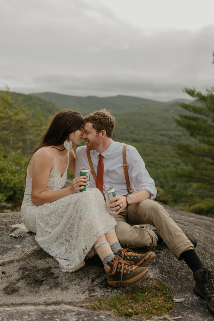 A bride kisses her groom on the nose on a mountain in North Carolina for their forest elopement. They are sitting down holding beers.