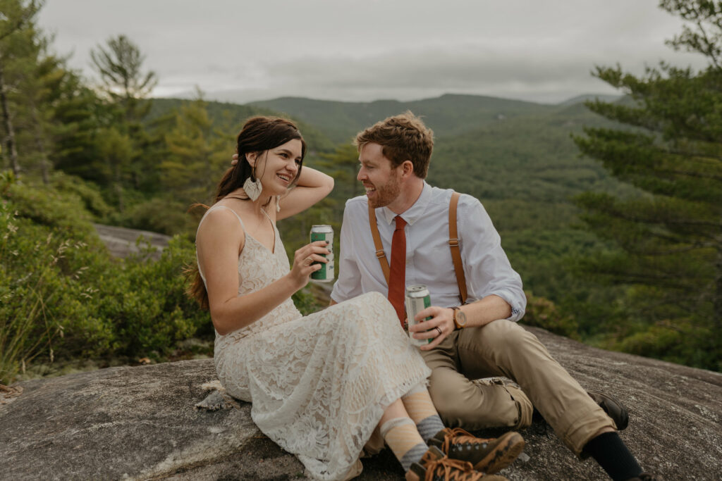 A bride and groom sit on a rock in North Carolina smiling at each other about to drink their special elopement beer.