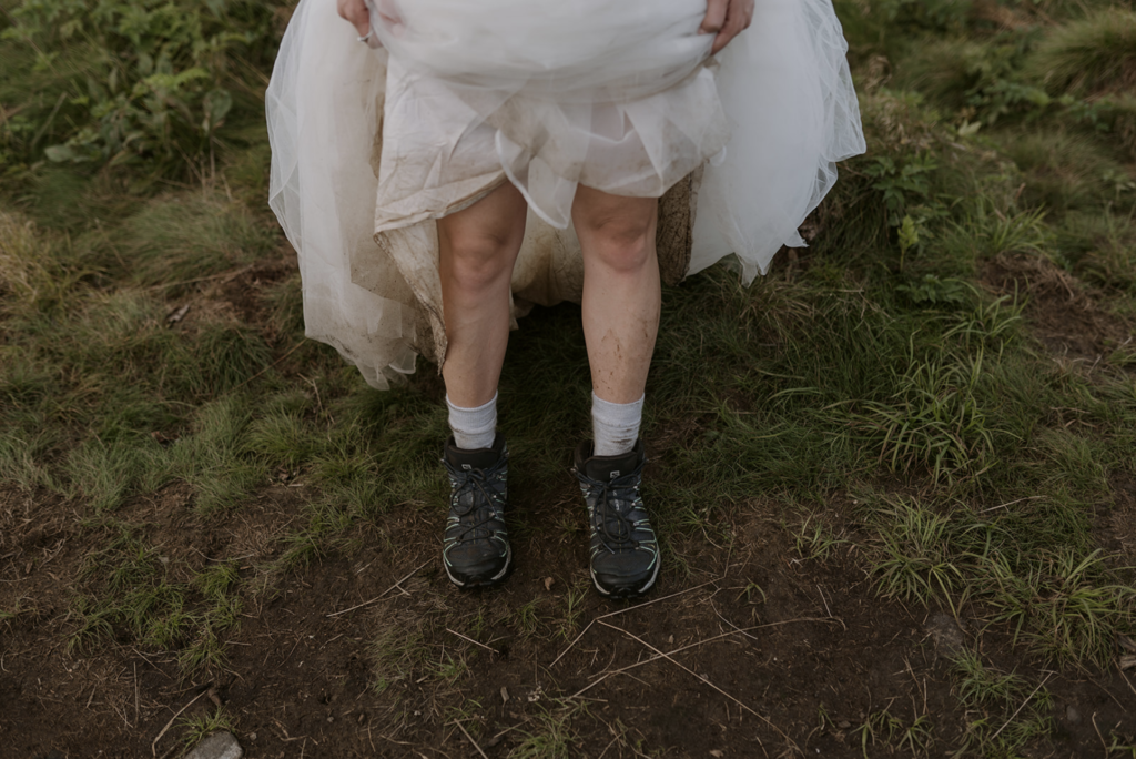 A bride is holding up the bottom of her dress to show off her dirty legs and hiking boots during her adventure elopement.