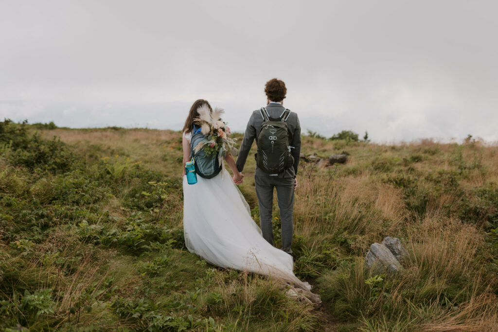 A couple stands holding hands in their wedding clothes on a mountain with a foggy view during their adventure elopement. The both have hiking backpacks on.