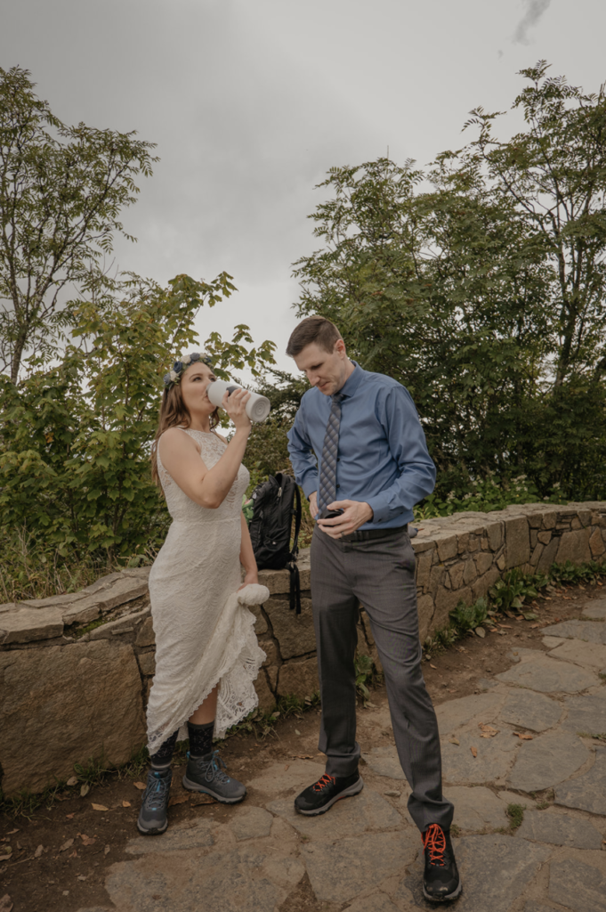 A couple is standing at a trail in their wedding clothes during an adventure elopement and the bride is drinking water while the groom holds the lid waiting for her.