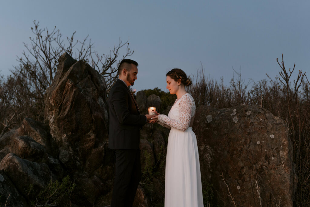 A couple on their elopement day in NC stands holding a single candle at dusk with their eyes closed.