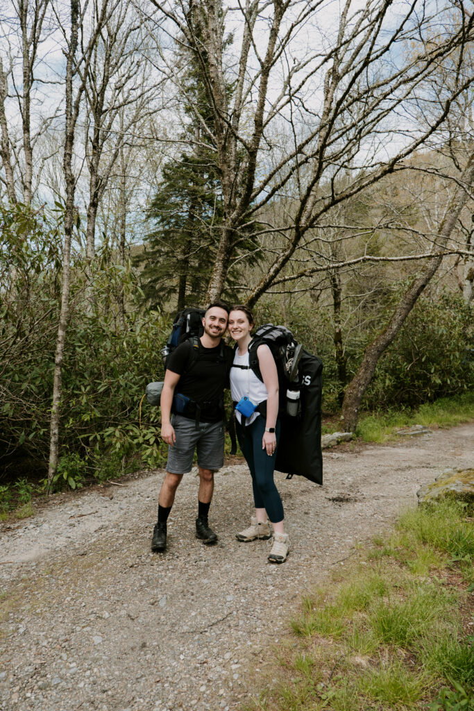 A couple stands smiling at the camera at the beginning of their hike for their elopement in NC. They have backpacks on to hike to a mountain and camp.