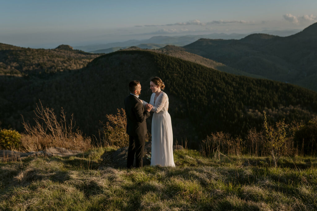 A bride and groom stand on a mountaintop in the blue ridge mountains during their backpacking elopement. The bride is helping the groom tie his tie.