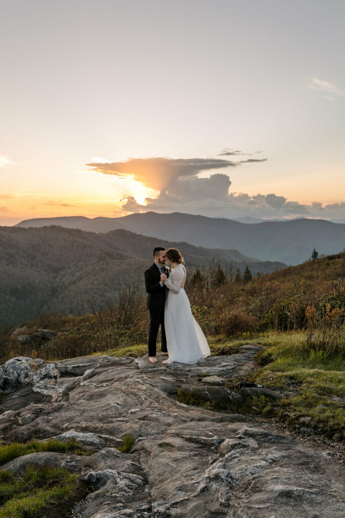 A couple stands in the distance on a mountain in NC for their elopements first dance on an open rock face at sunset.