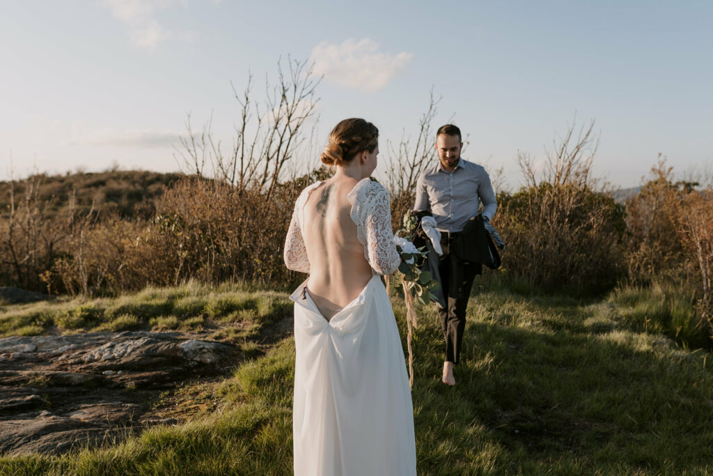 A bride is walking towards a groom with her dress unzipped. They are on a mountaintop and they are getting ready for their elopement ceremony.