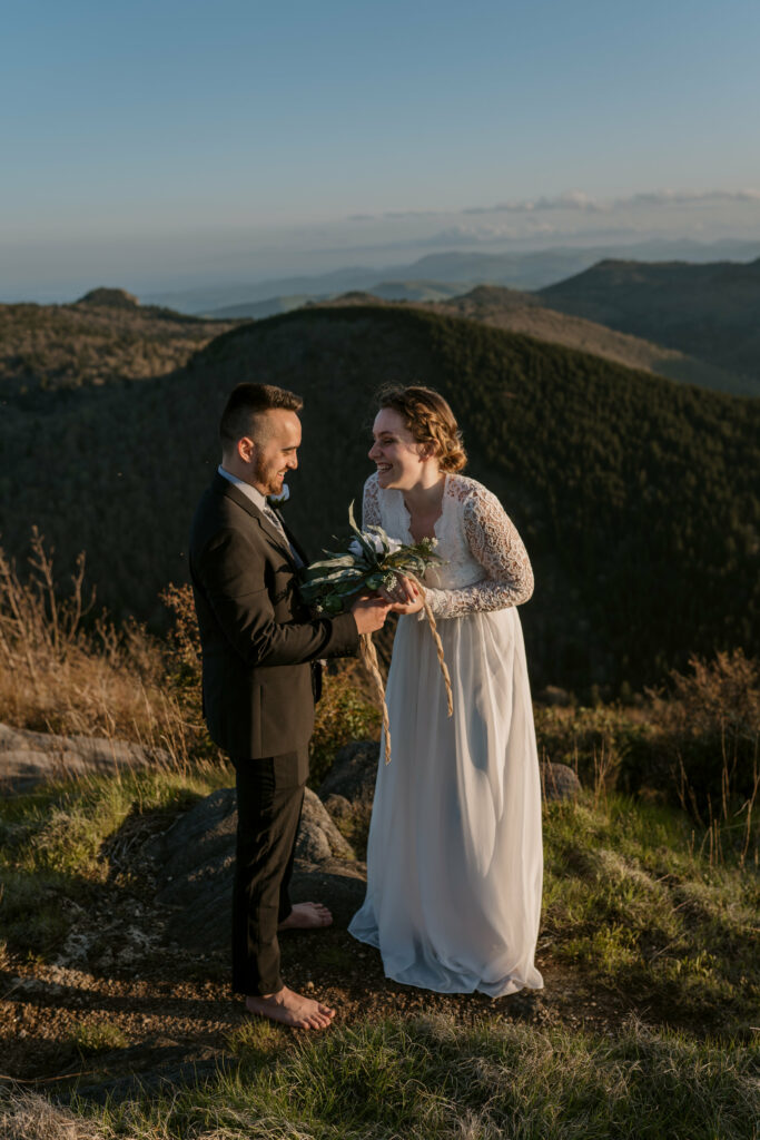 A bride and groom stand giggling as the groom helps put on her floral wristlet. They are in the blue ridge mountains of NC for their elopement.
