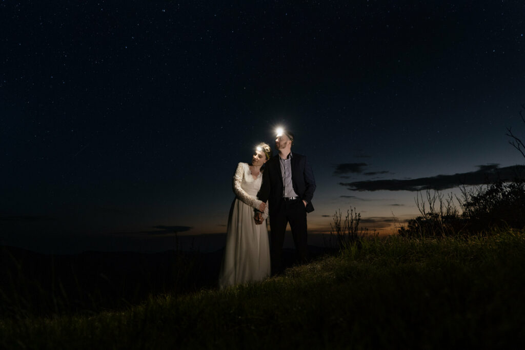 A couple stands side by side looking into the distance with headlamps lighting them up during sunset on their elopement day in NC.