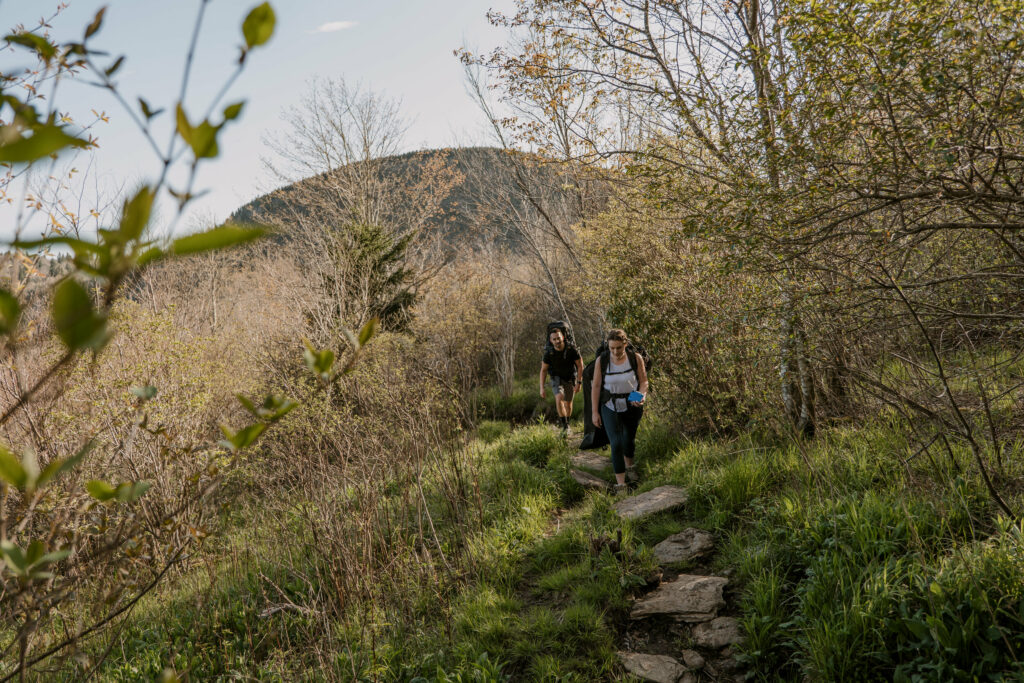 A couple backpacks up a grassy trail with a small view of the blue ridge mountains in the background in NC