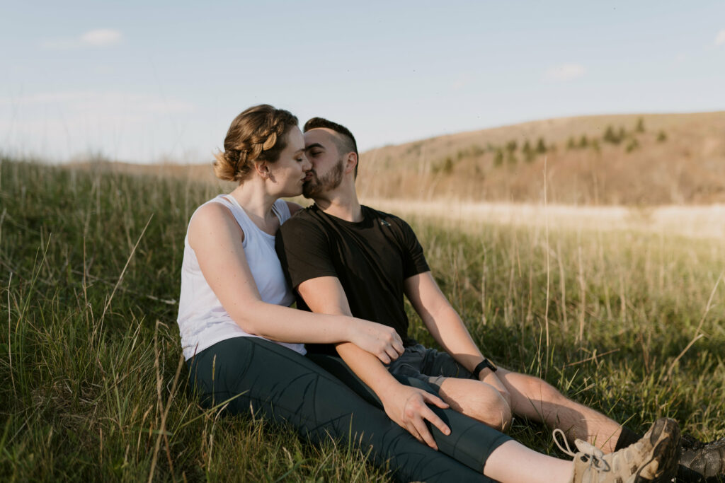A couple sits side by side kissing in a tall grass field.