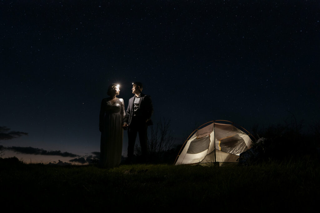 A couple stands side by side facing each other beside their a lit up tent for their backpacking elopement in NC. The sun has set and there are stars in the sky.