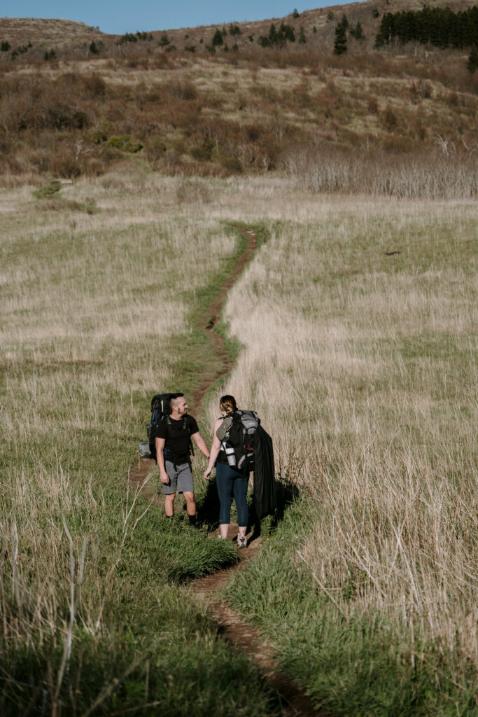 A couple stands on a trail surrounded by tall dead grass with their backpacks on, looking in to the distance.