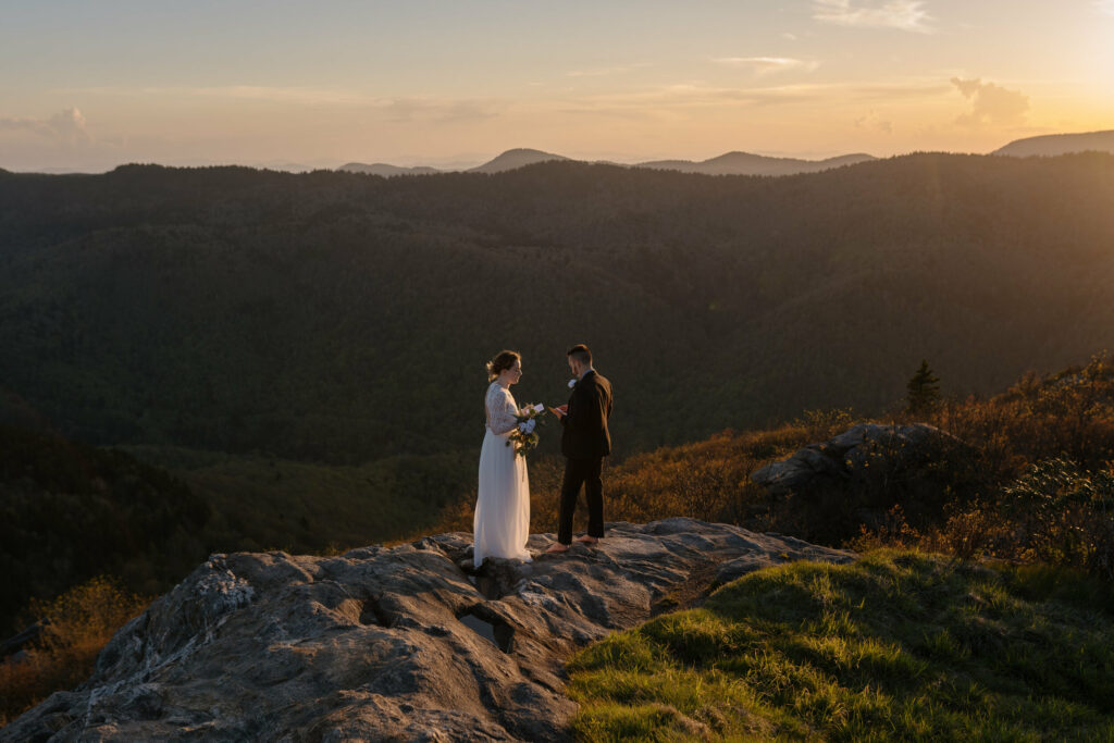 A bride and groom stand facing each other and holding their vow books on a rock at sunset during their backpacking elopement in NC. There are big blue ridge mountains in the background.