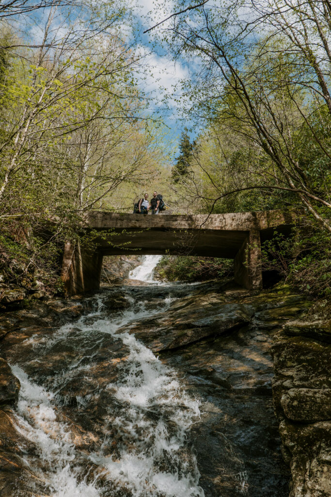 A couple stands in the distance high on a bridge crossing a waterfall during their backpacking elopement in NC.