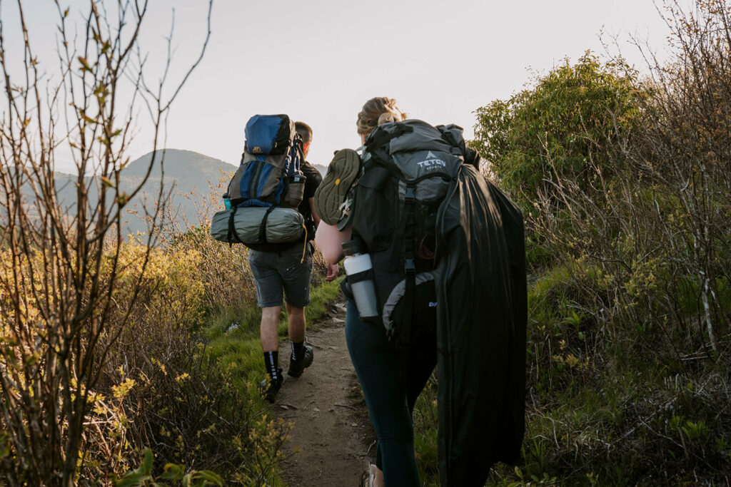 A couple is hiking with backpacks on on a trail through the mountains - the bride has a clothing back strapped to her pack with all their wedding clothes inside. They are backpacking to their elopement location.